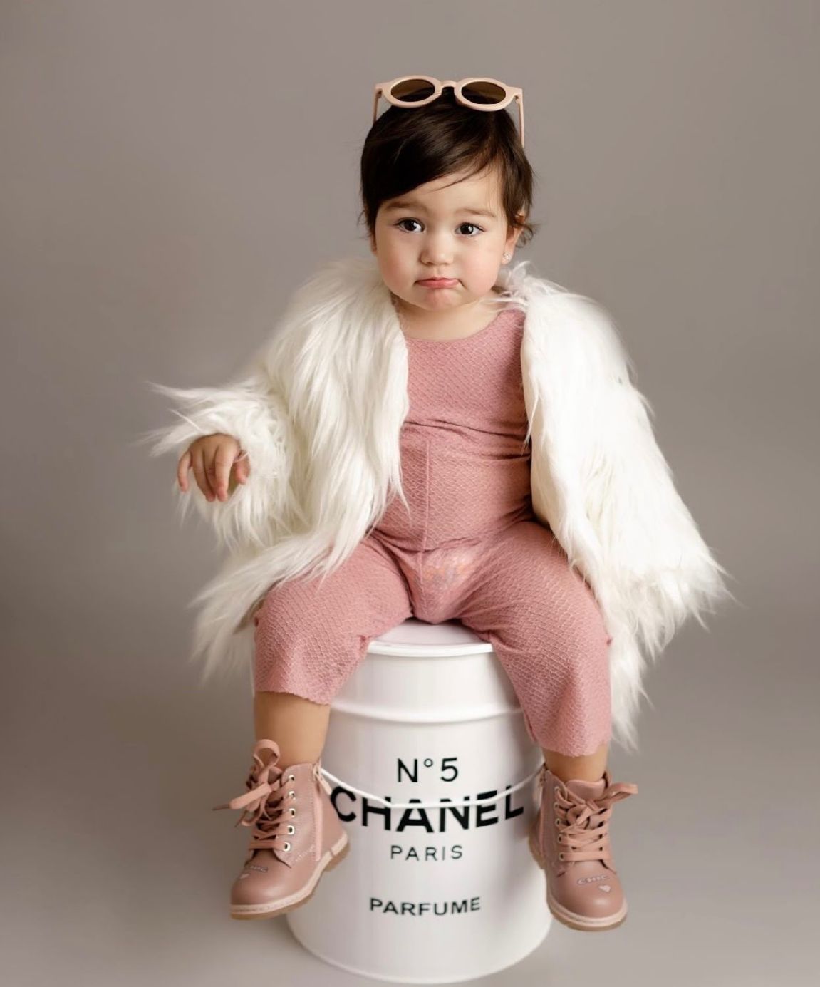 Stylish toddler in pink jumpsuit and white fur jacket poses on Chanel No 5 decorative container wearing sunglasses on head.