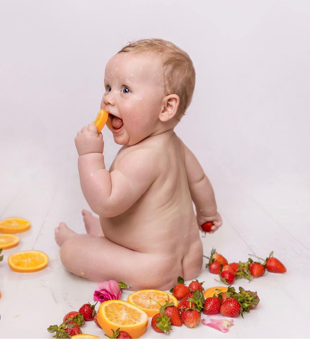 Baby sitting on white background surrounded by fresh oranges and strawberries in artistic food photography setup.