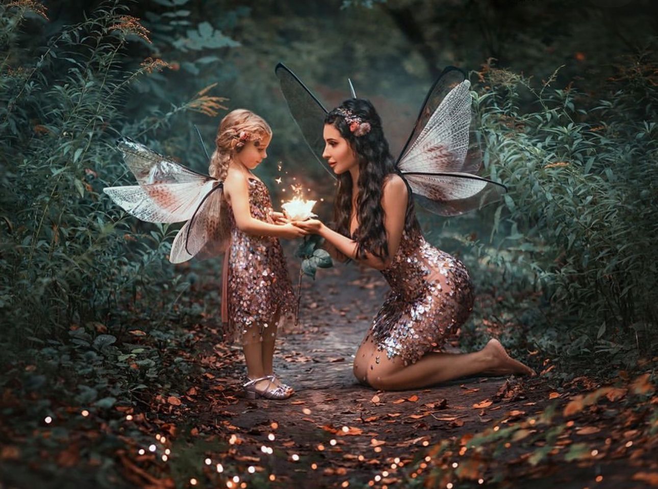 Two fairy women in sequin dresses with wings share magical glowing light in an enchanted forest setting.