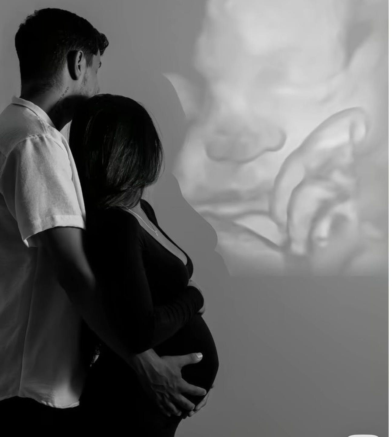 Black and white photo of pregnant couple embracing with ultrasound image projected on wall behind them.