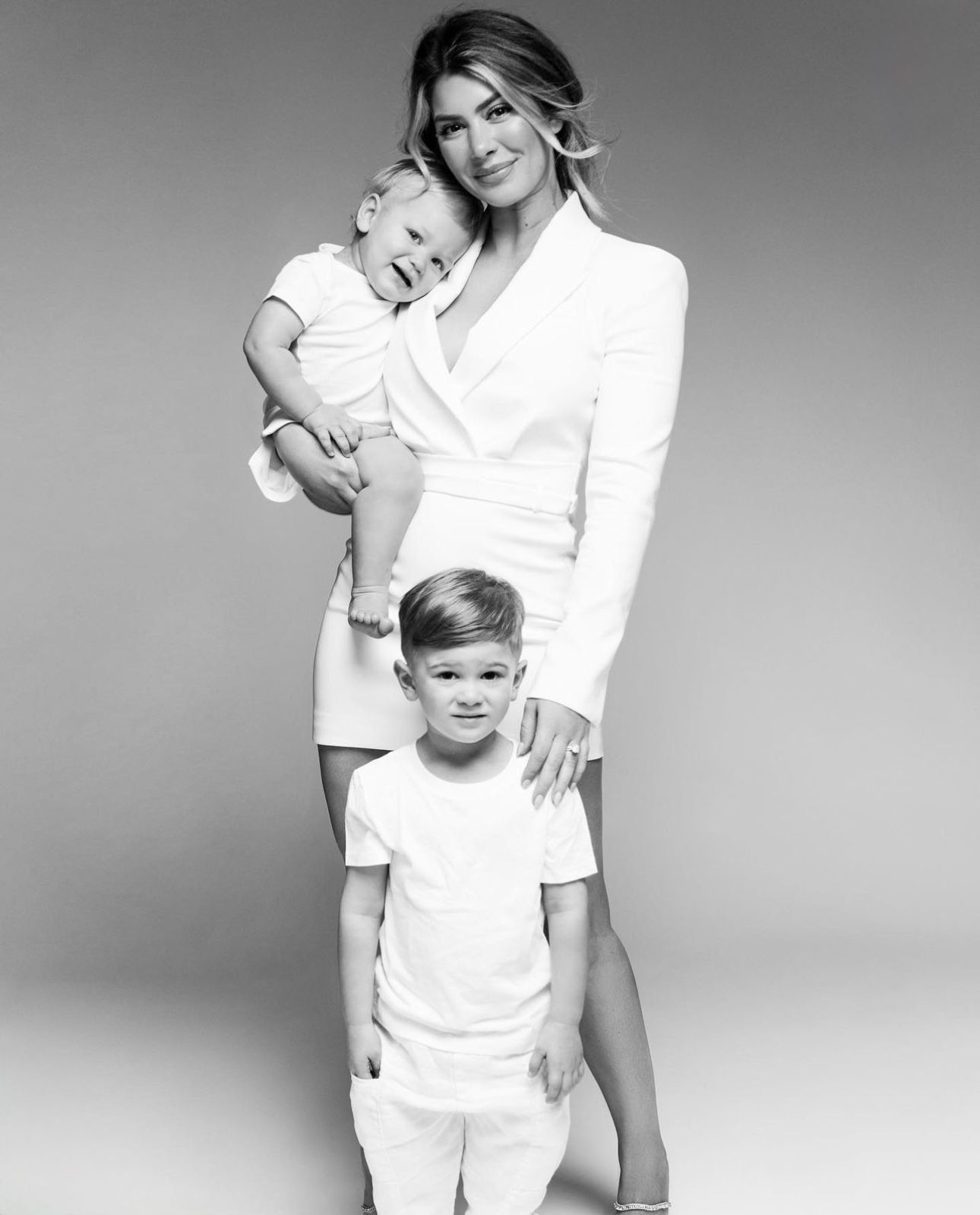 Black and white photo of a woman in white dress holding a toddler, with a young boy standing in front.
