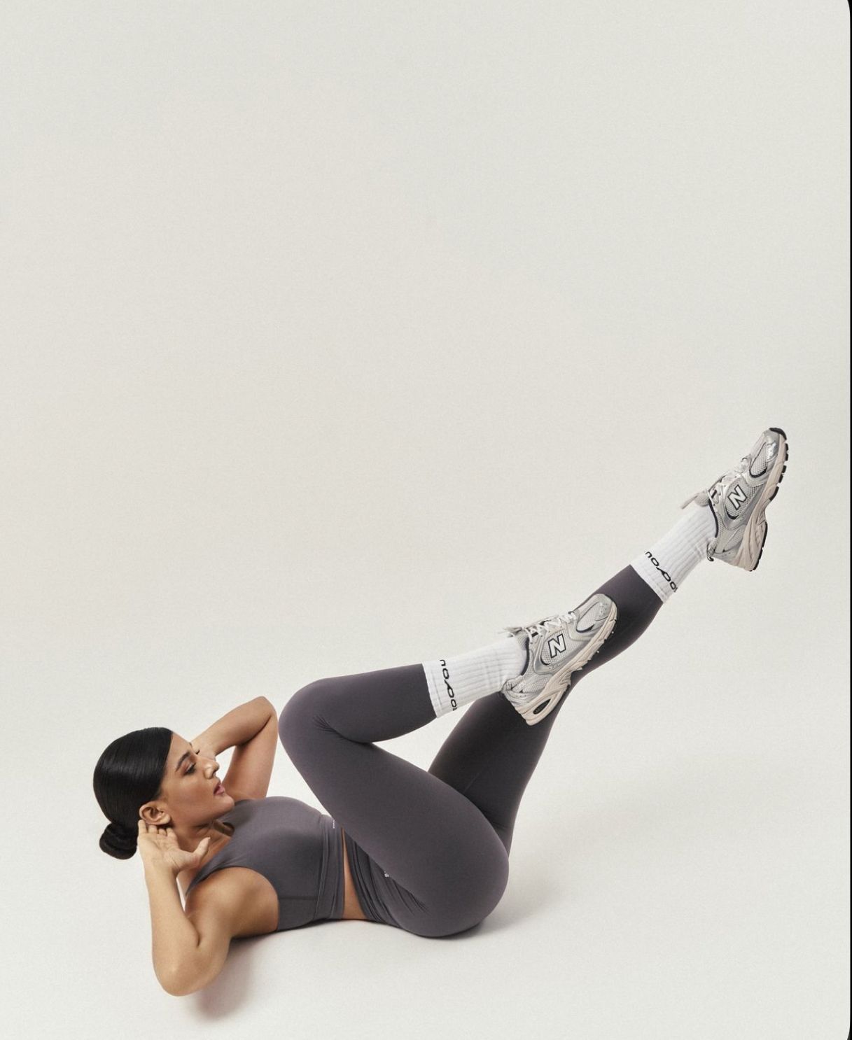 Woman in gray athletic wear doing bicycle crunches exercise on white background wearing white socks and sneakers.
