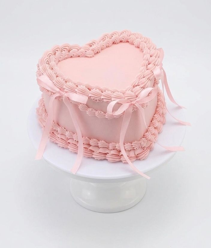 Pink heart-shaped cake with piped frosting borders and satin ribbon bows on a white cake stand.