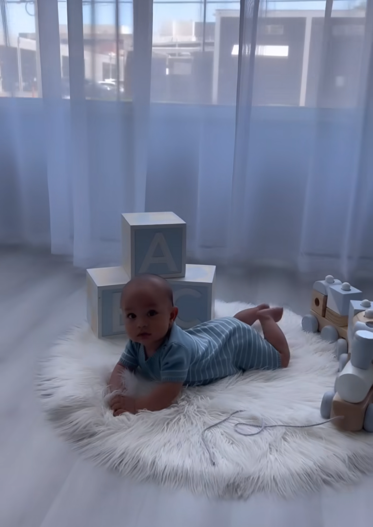 Baby doing tummy time on fluffy white rug surrounded by wooden alphabet blocks and toys near sheer curtains.