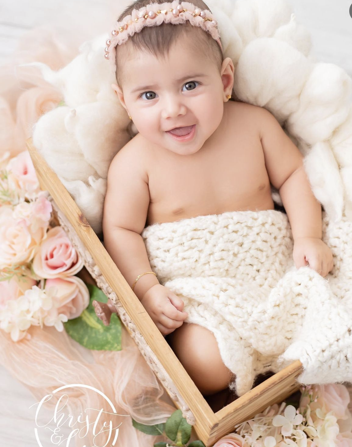Smiling baby girl in a wooden crate with a cream knit blanket, floral headband, and pink roses in a professional photo.