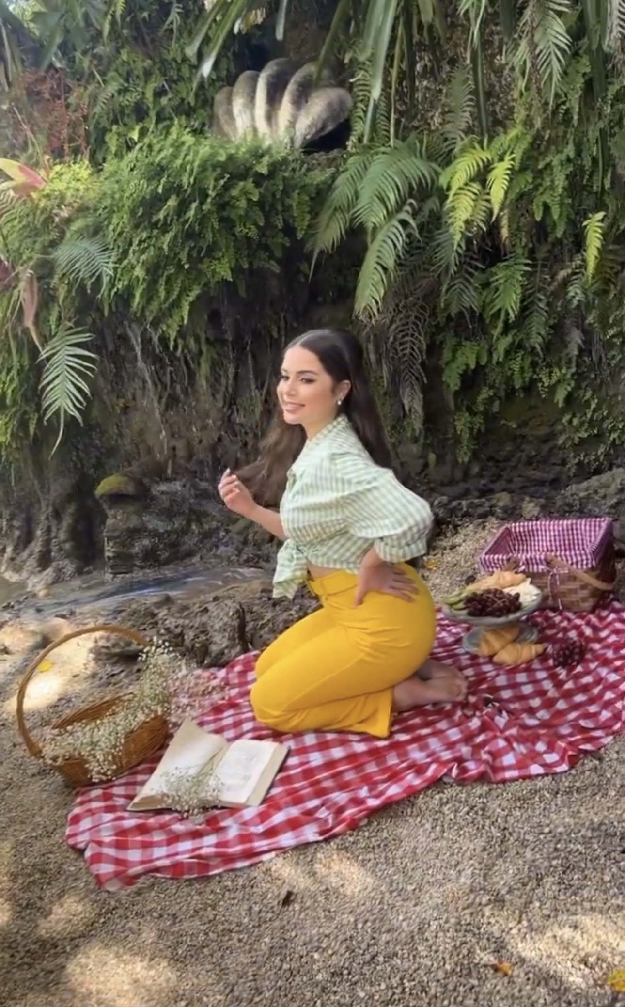 Woman in yellow pants and striped top enjoying a picnic on red gingham blanket surrounded by tropical greenery.