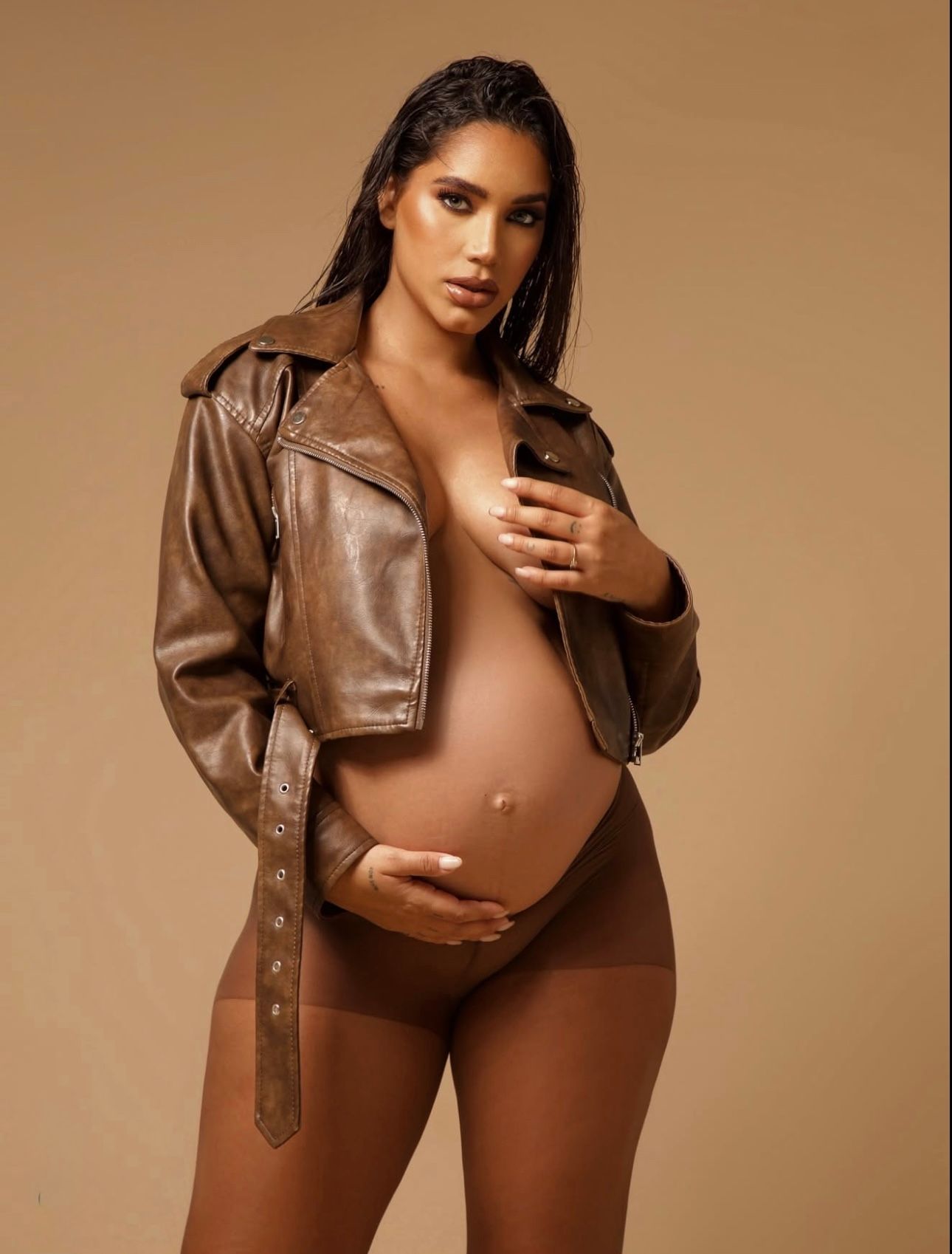 Pregnant woman in brown leather jacket posing for maternity photoshoot against neutral background.