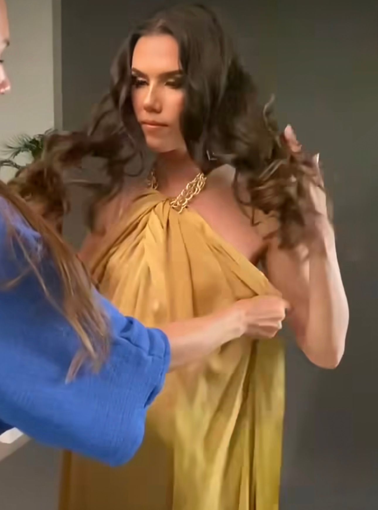 Woman in gold halter dress with chain necklace gets styling assistance from person in blue top.