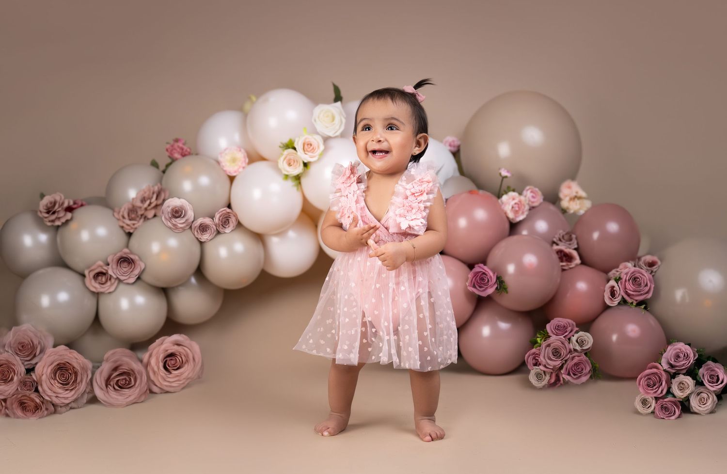 Baby girl in pink dress stands smiling before balloon and rose backdrop at first birthday cake smash photo session.