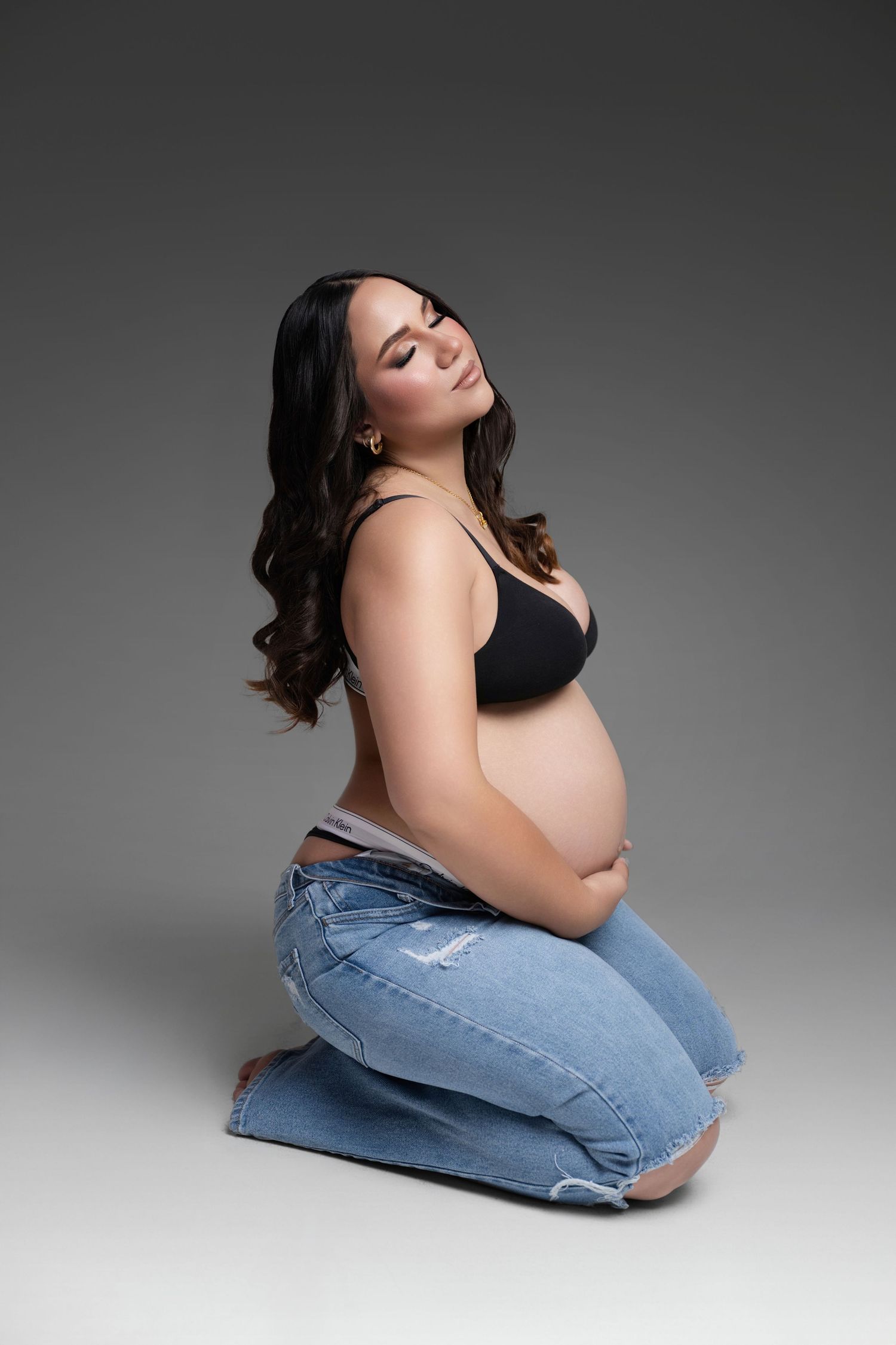 Pregnant woman in black bra and ripped jeans kneeling on studio floor, cradling baby bump, smiling softly.
