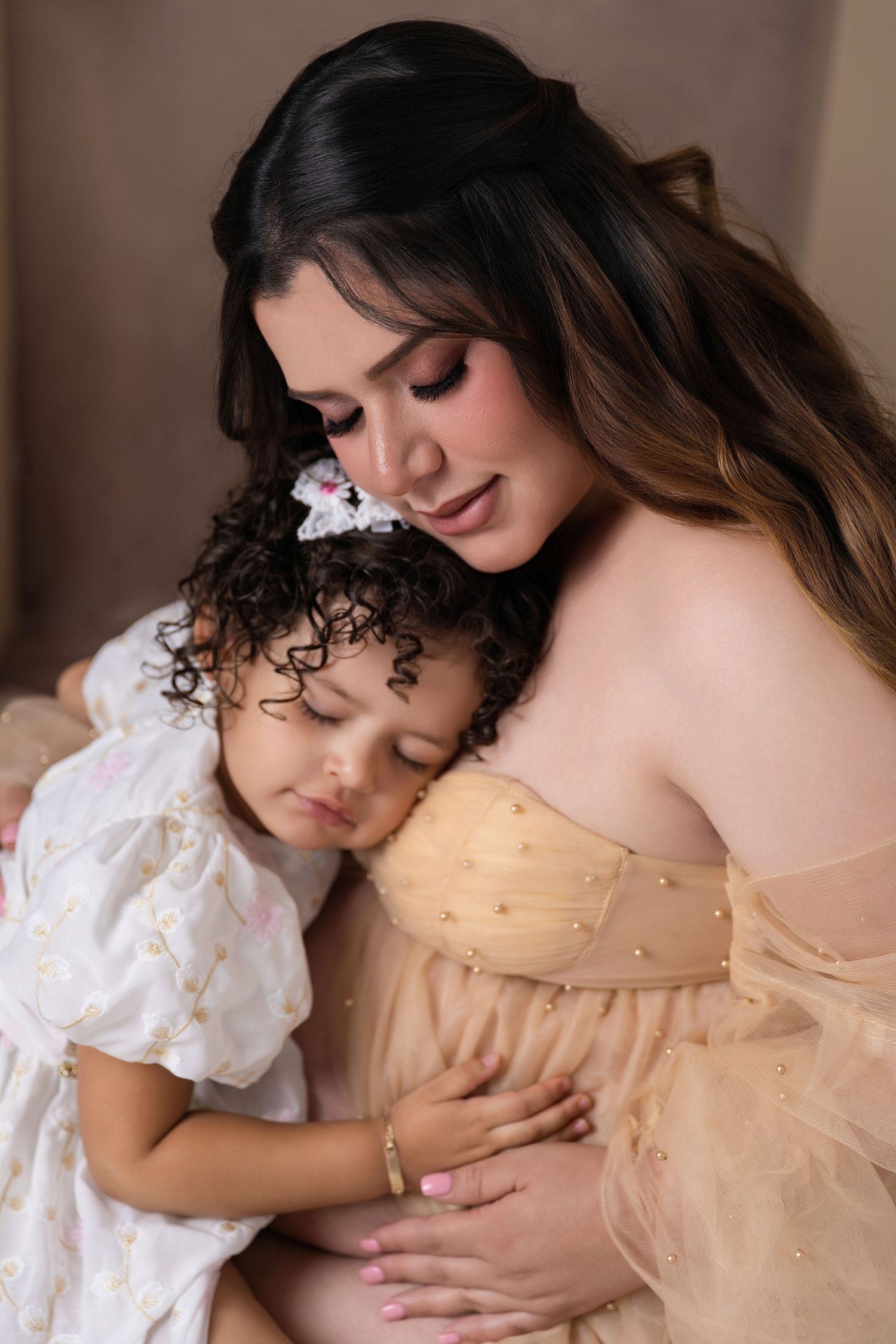 A woman in a beige tulle gown tenderly embraces a curly-haired toddler girl in a white floral dress.