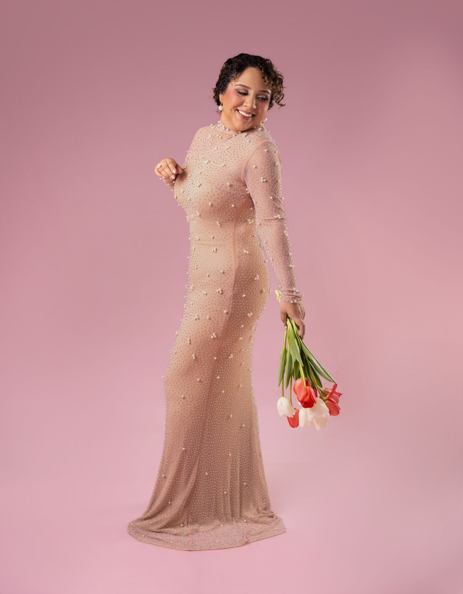 Smiling woman in a long sleeved blush gown with pearl embellishments holds a bouquet of red and white tulips against a pink studio backdrop.