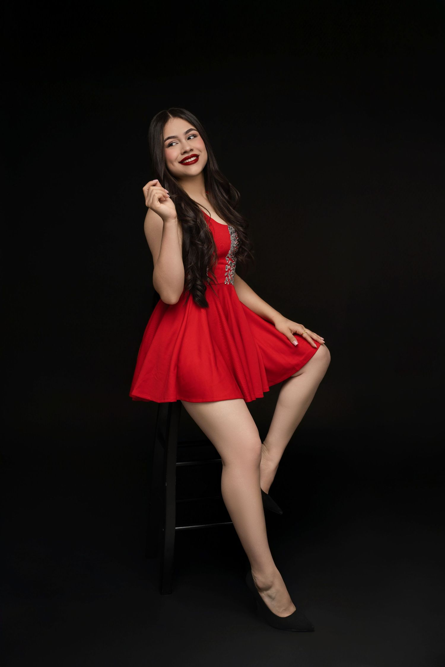 Woman with long dark wavy hair in a red mini dress and black heels poses on a stool against a black studio background.