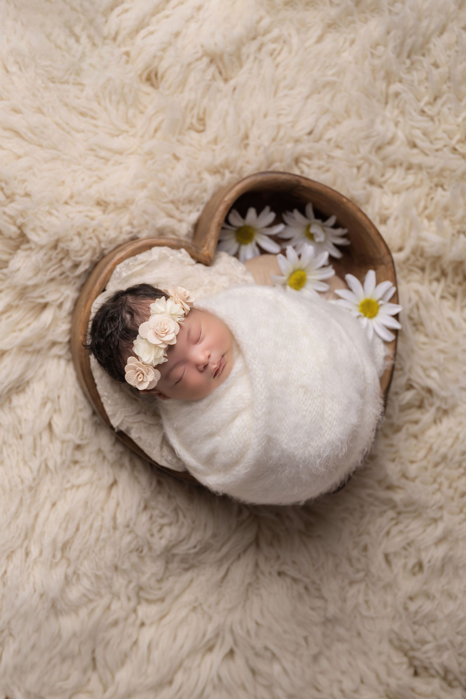 Newborn baby girl swaddled in white wrap, wearing floral headband, sleeping in heart-shaped wooden bowl with daisies.