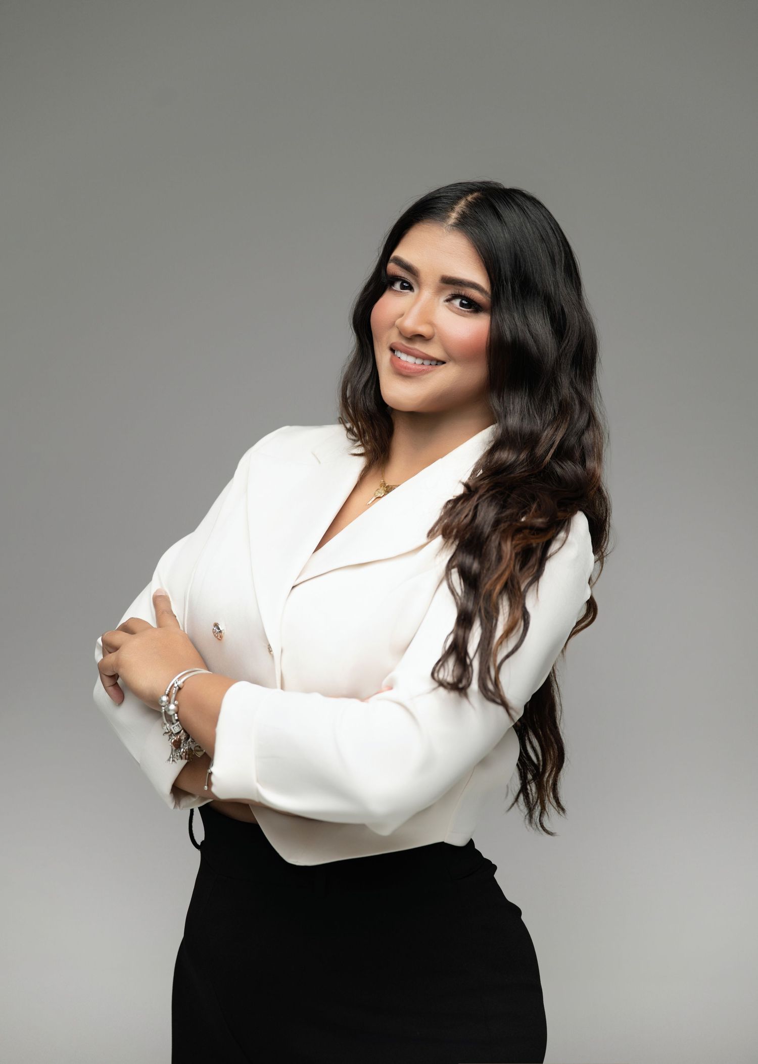 Professional woman in white blazer and black skirt poses confidently with arms crossed against gray background.