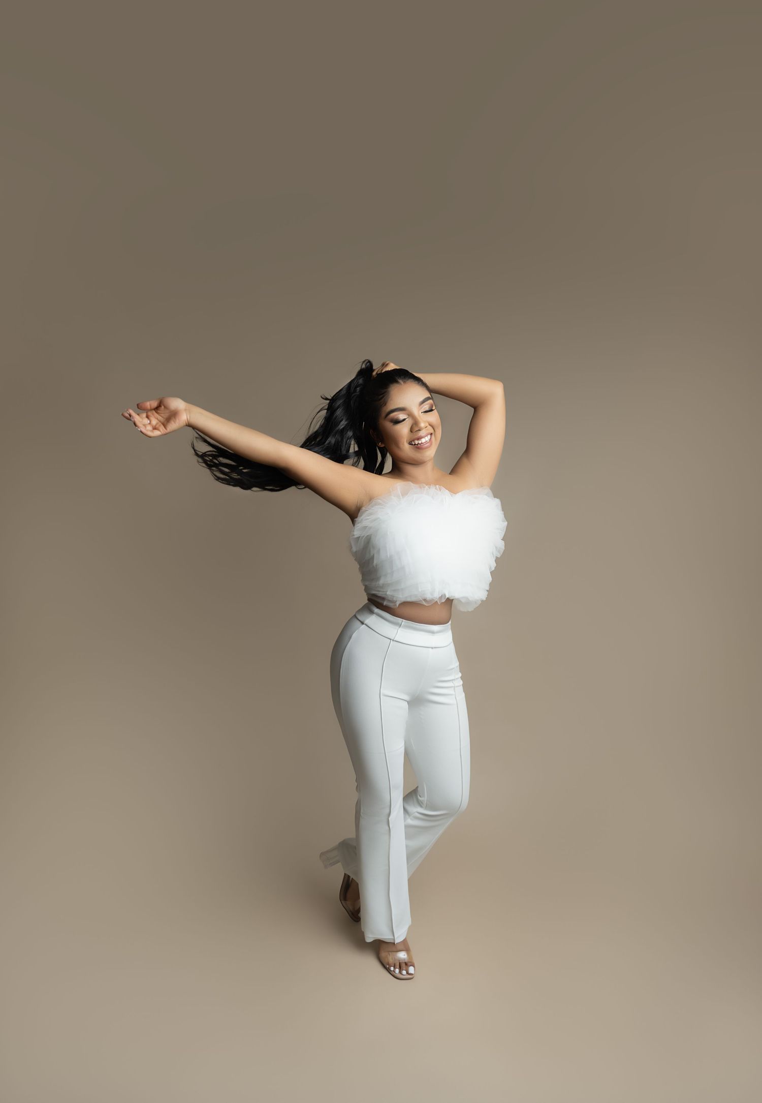 Woman with long dark hair smiles and dances in a white fluffy tulle top and white flared pants against a neutral beige studio backdrop.