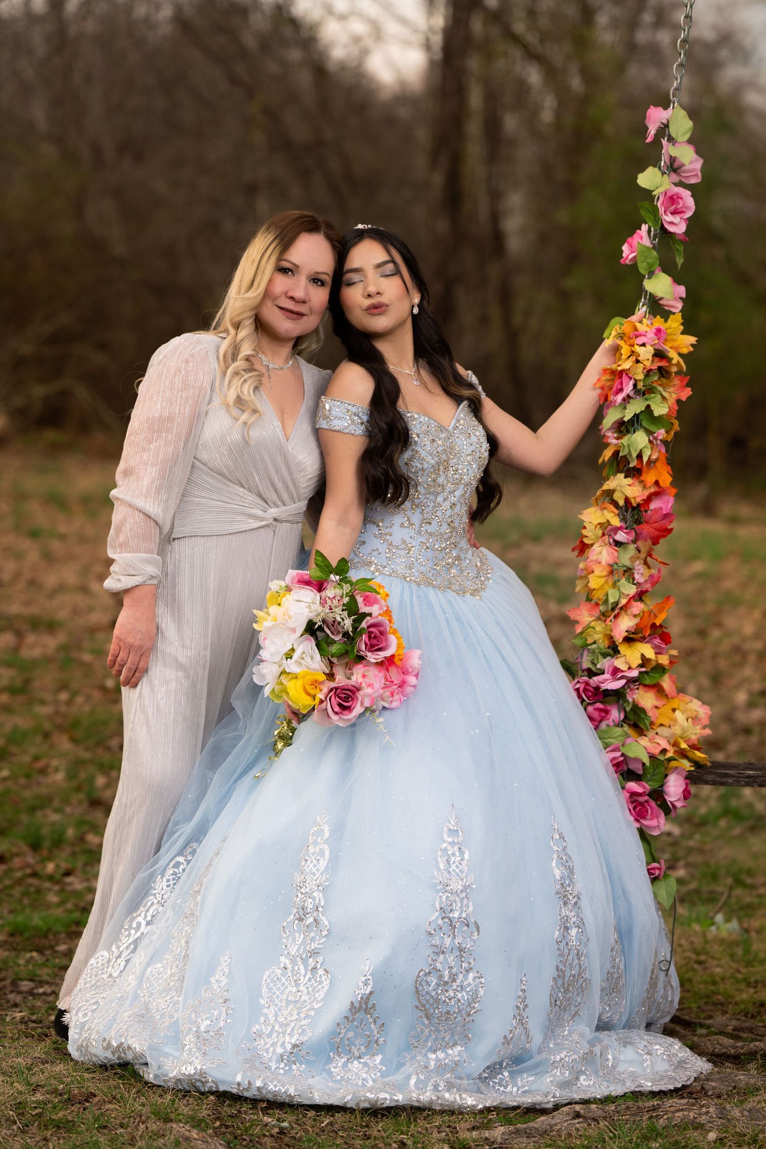 Two women in elegant dresses pose by a floral swing outdoors, one in a blue quinceañera gown holding a colorful bouquet.