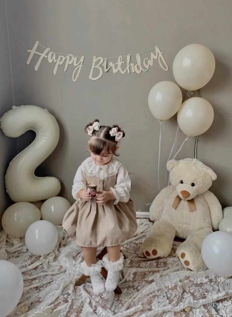 A toddler girl celebrates her 2nd birthday with balloons, a giant teddy bear, and a Happy Birthday banner.