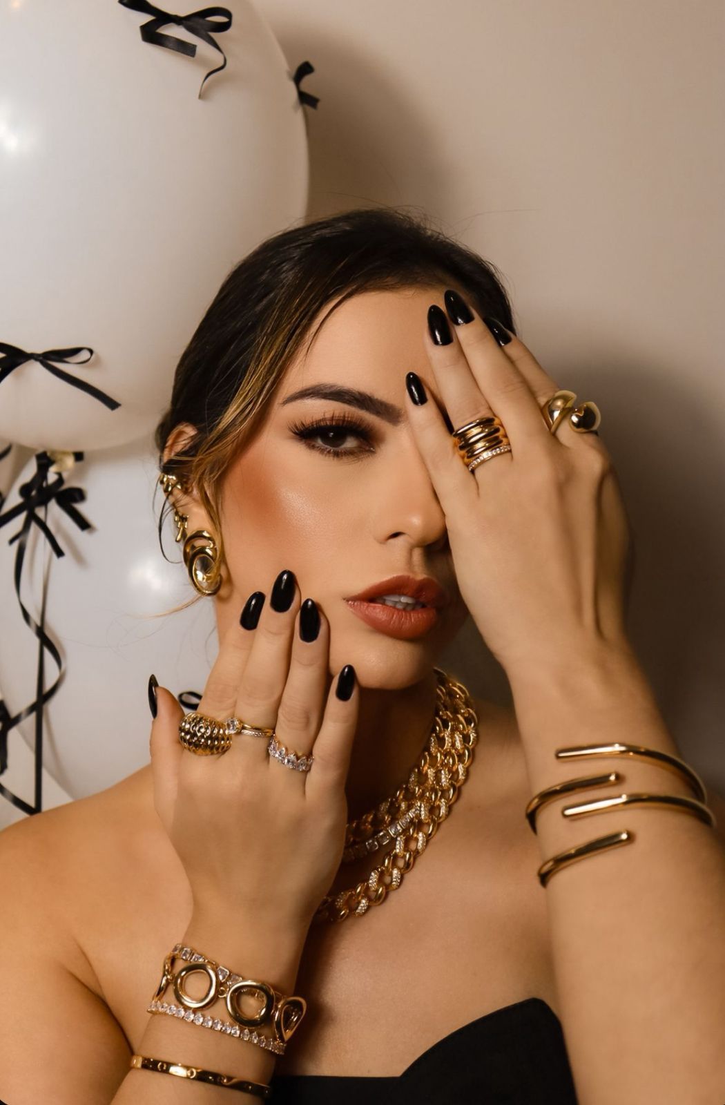 Woman posing with gold jewelry, rings, bracelets, and chain necklace, black nails, near white balloons.