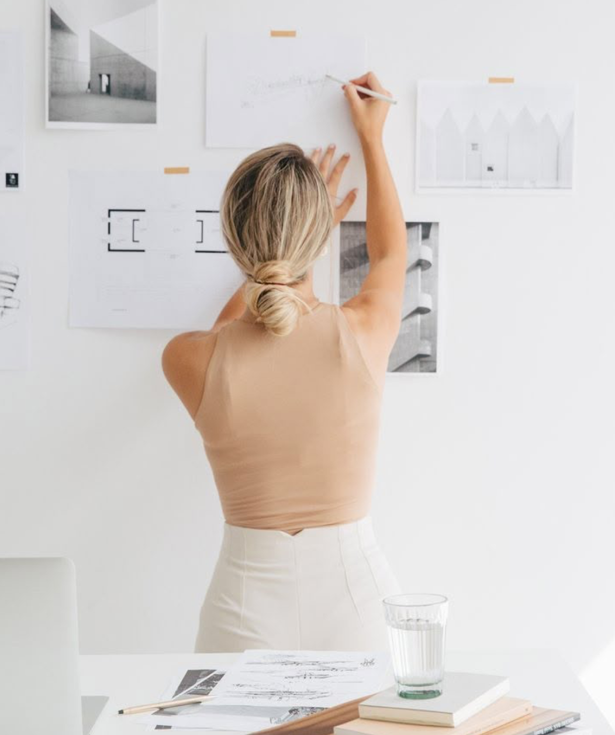 Woman with blonde hair in a bun writing on architectural sketches pinned to a white wall in a bright studio.