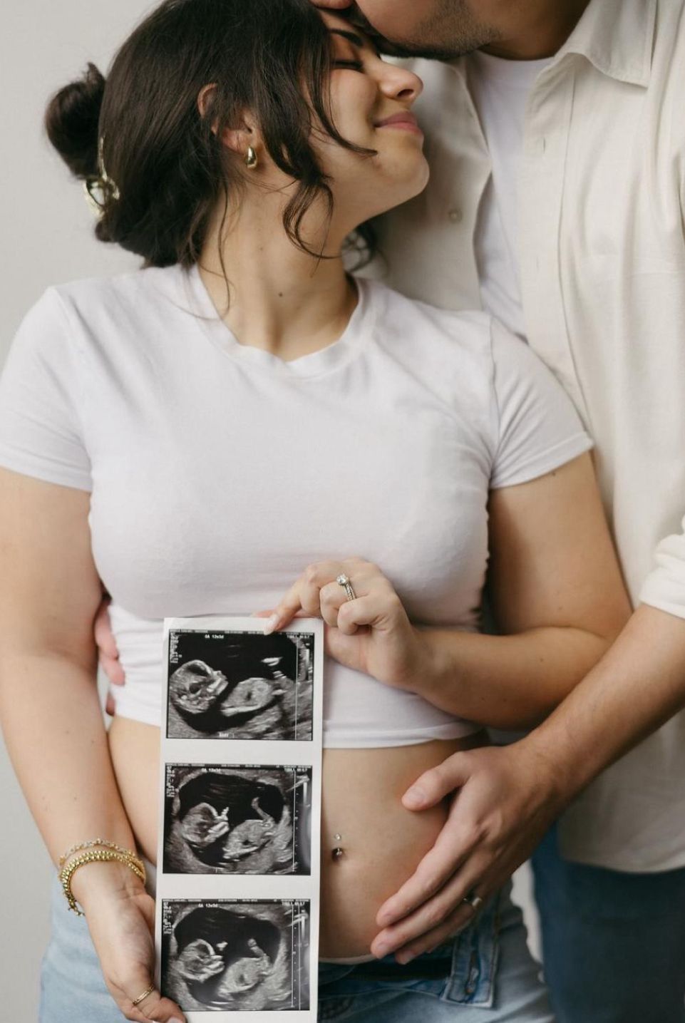 Pregnant couple holding ultrasound photos, woman in white crop top with baby bump, partner kissing her cheek tenderly.