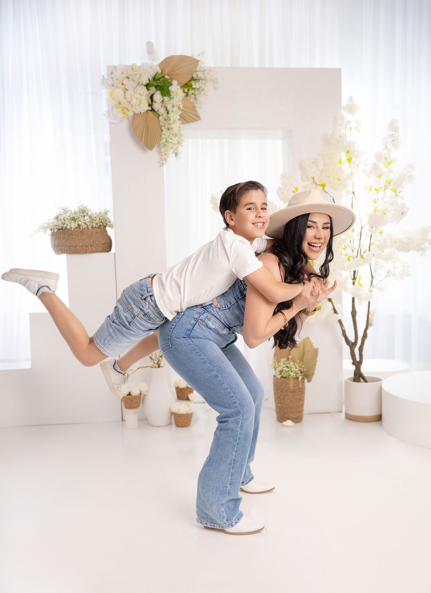 A woman in a hat gives a laughing child a piggyback ride in a bright white floral studio setting.