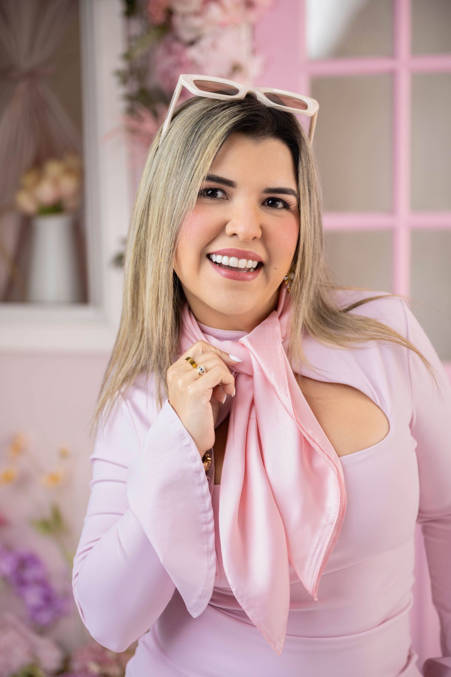 Smiling blonde woman in pink outfit with scarf and sunglasses on head, posing in front of floral pink backdrop.