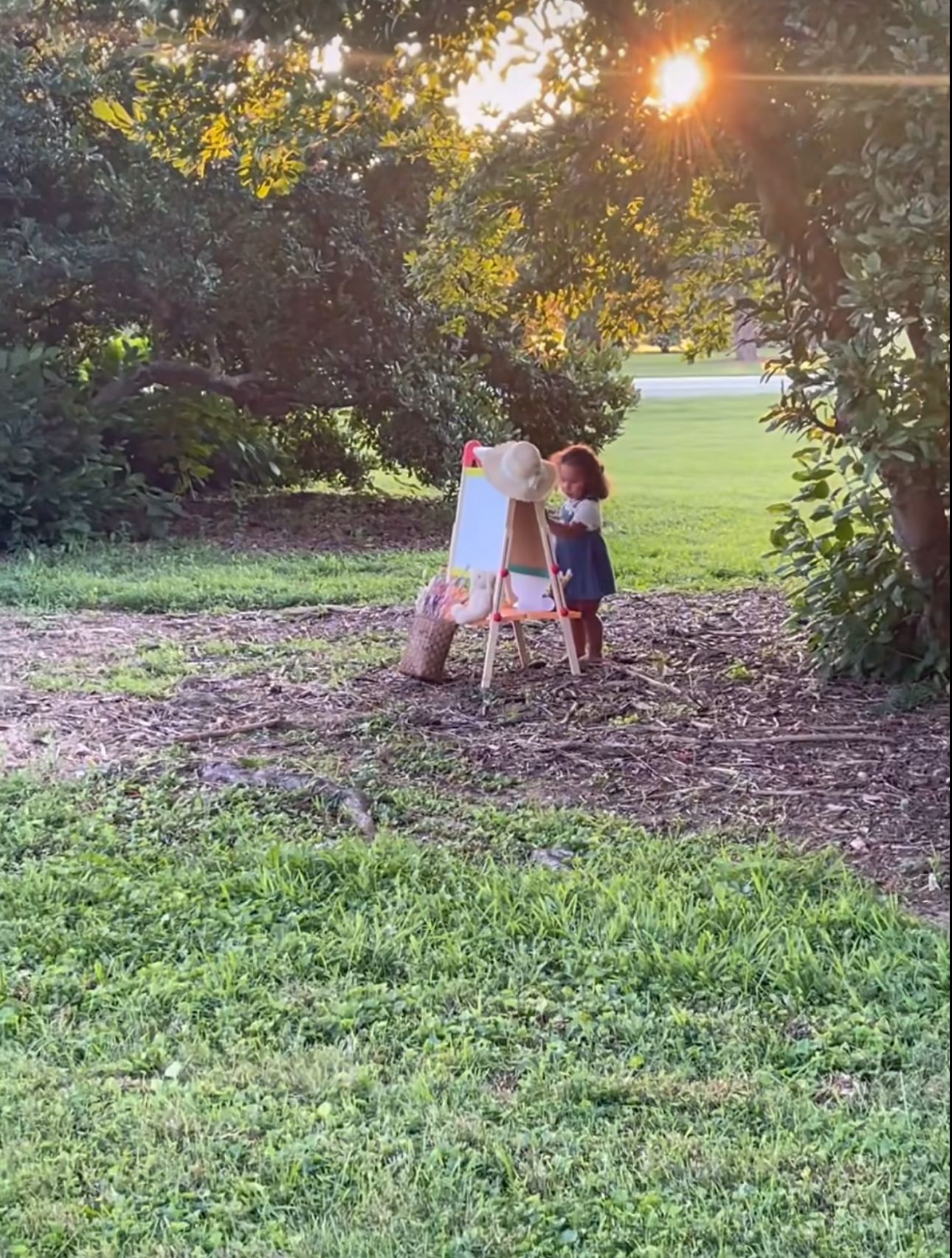 Young child painting at an easel outdoors under trees during a beautiful golden sunset.