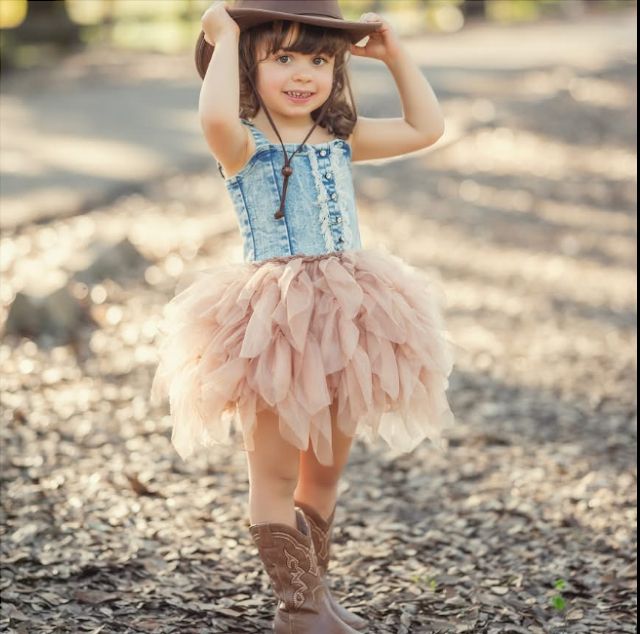 Adorable little girl in denim top, pink tutu skirt, cowboy boots and hat posing outdoors in golden sunlight.