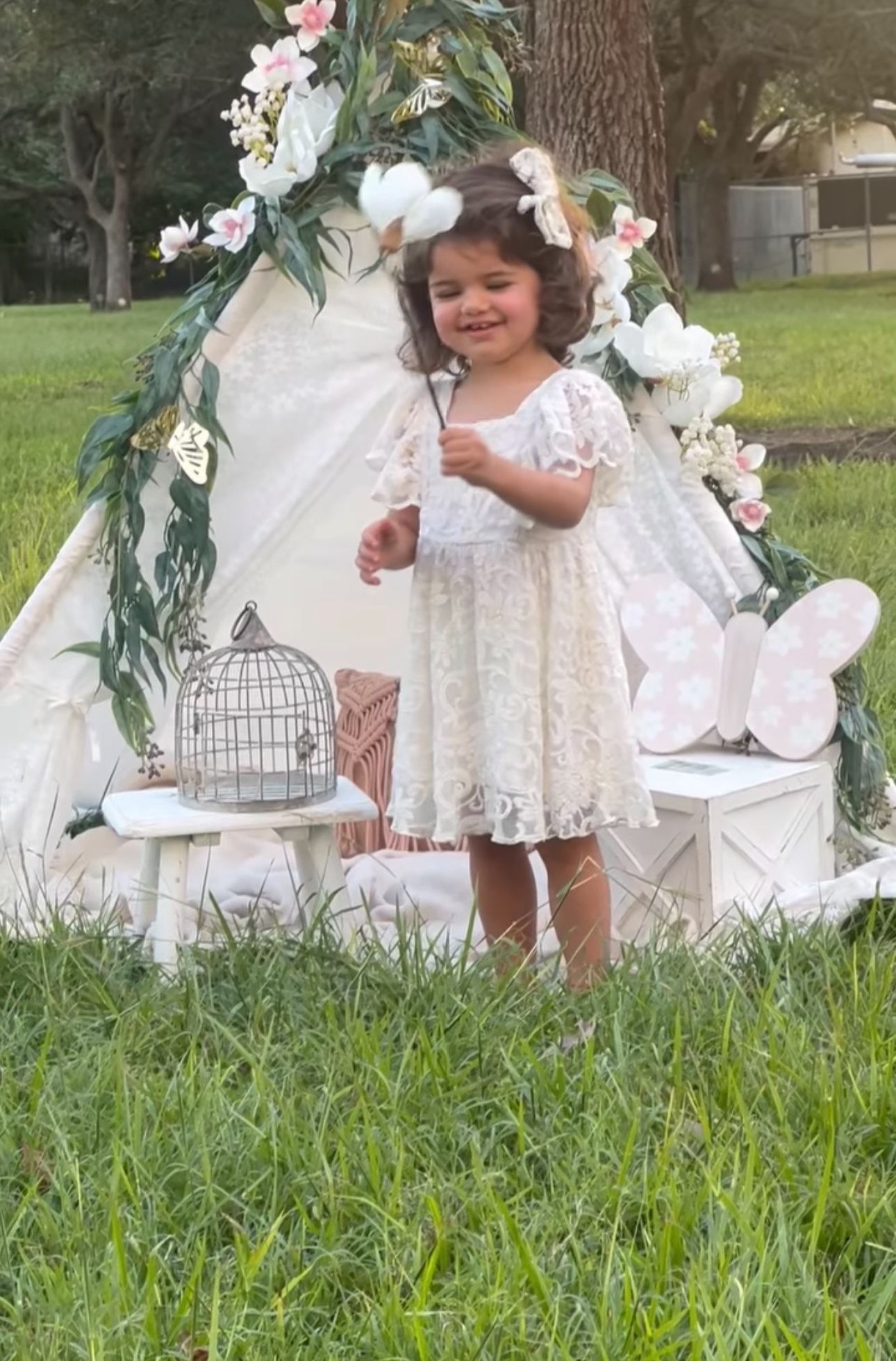 Adorable toddler girl in white lace dress stands before a floral teepee backdrop during an outdoor photoshoot.
