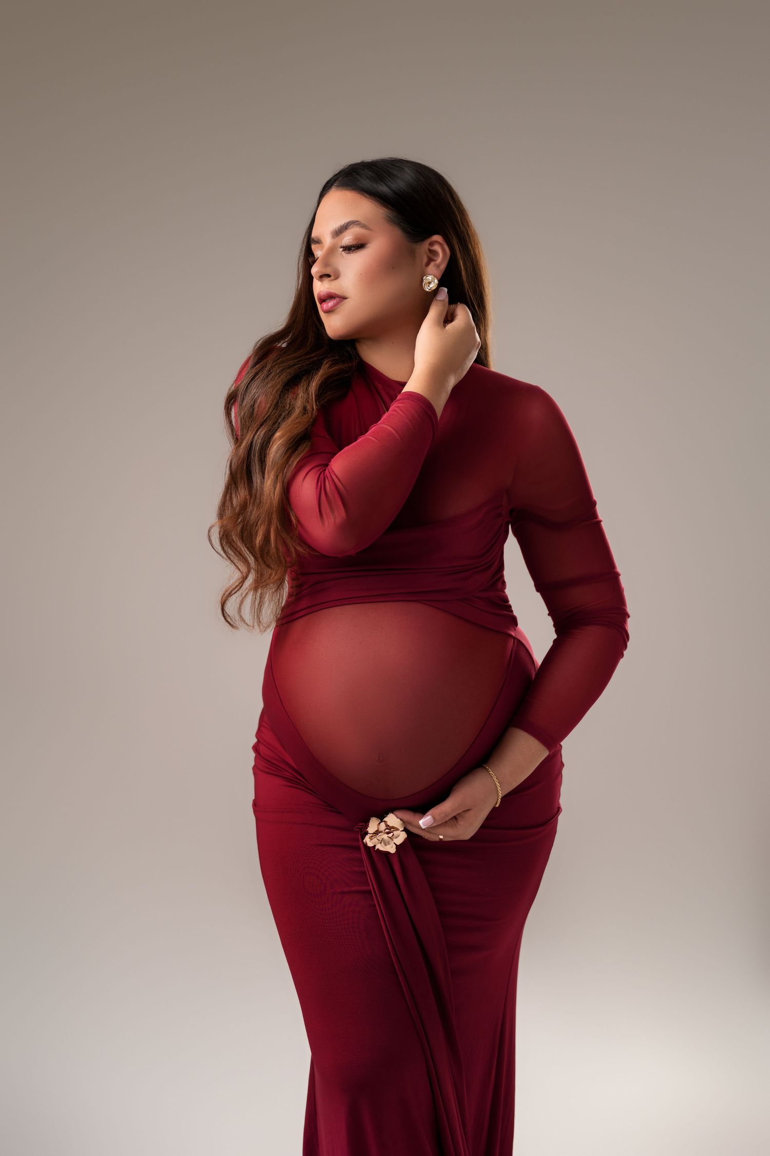 Pregnant woman in elegant red long-sleeve maternity gown posing gracefully against a neutral studio background.