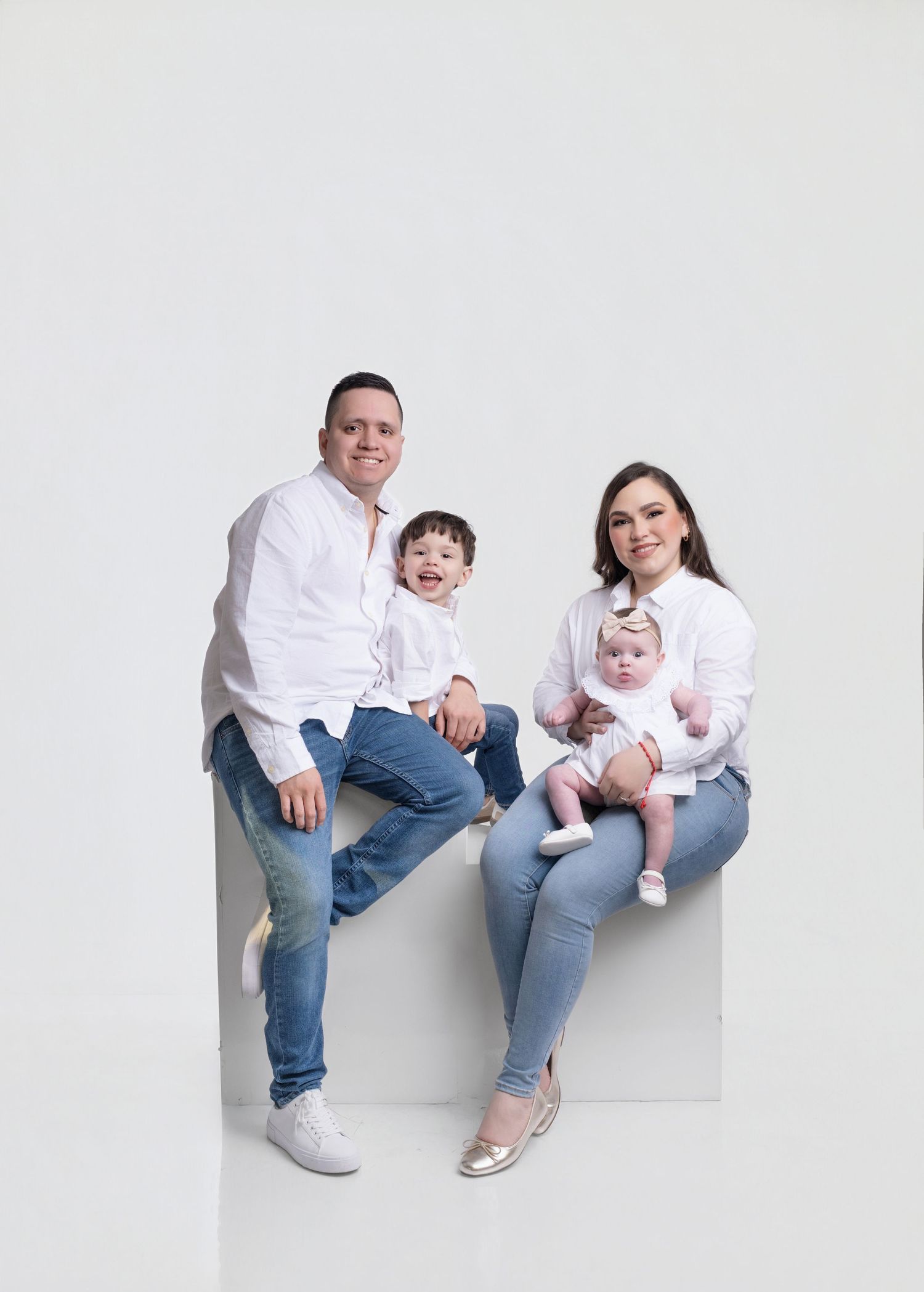 Family of four in matching white shirts and jeans posing for a studio portrait with two young children.