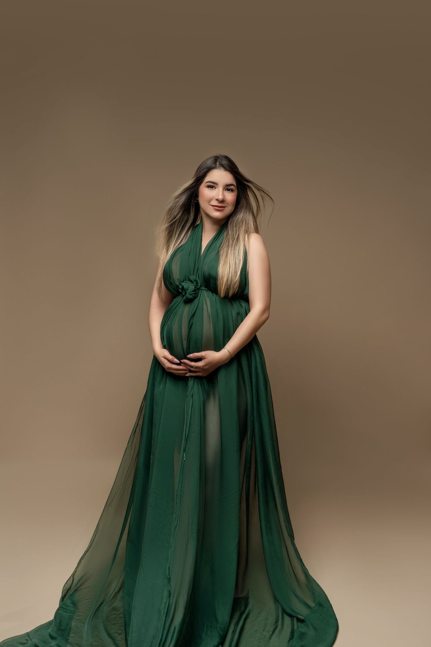 Pregnant woman in elegant green chiffon gown cradling baby bump in professional maternity studio portrait.