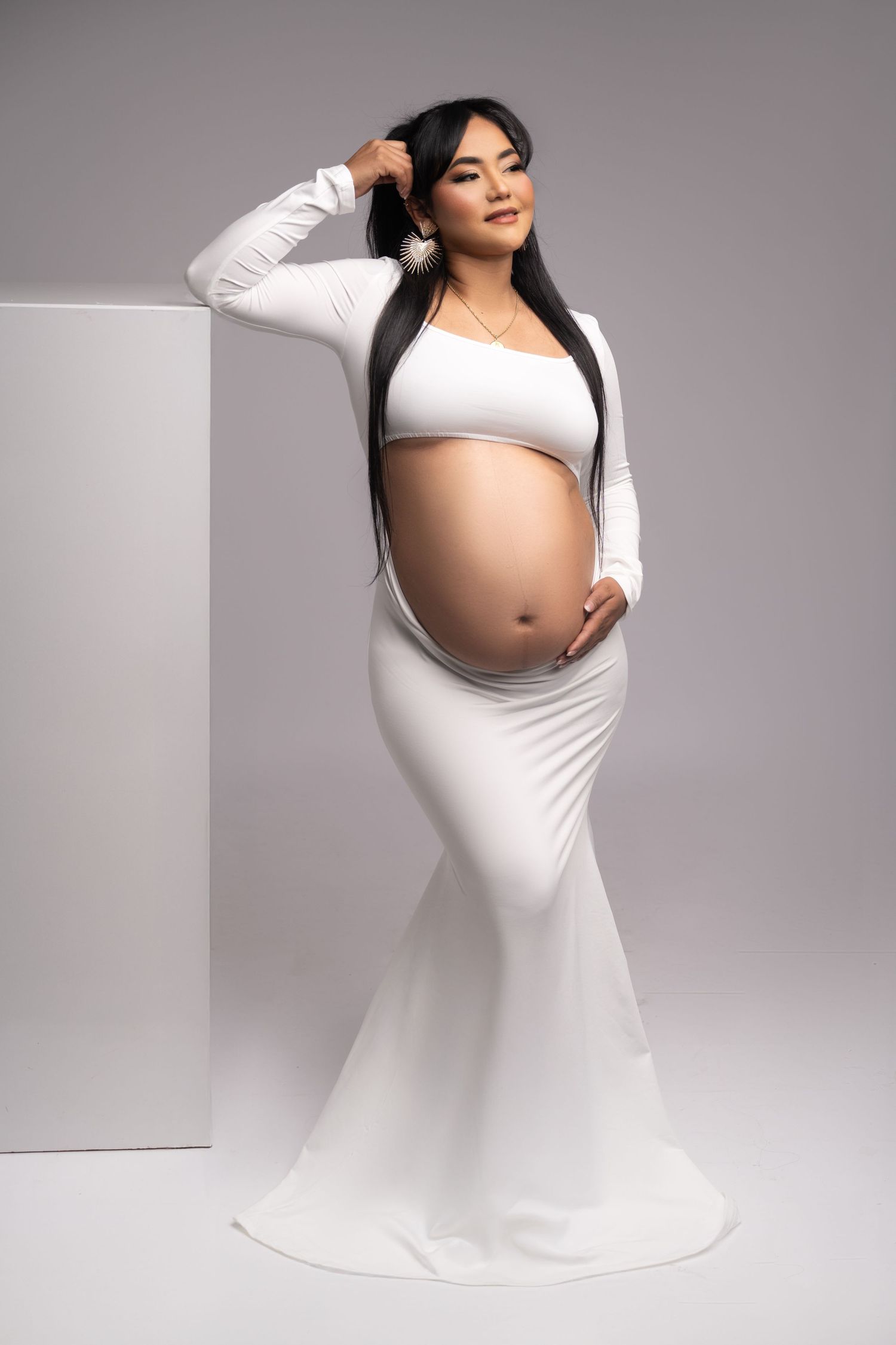 Professional maternity portrait in fitted white gown against grey backdrop.