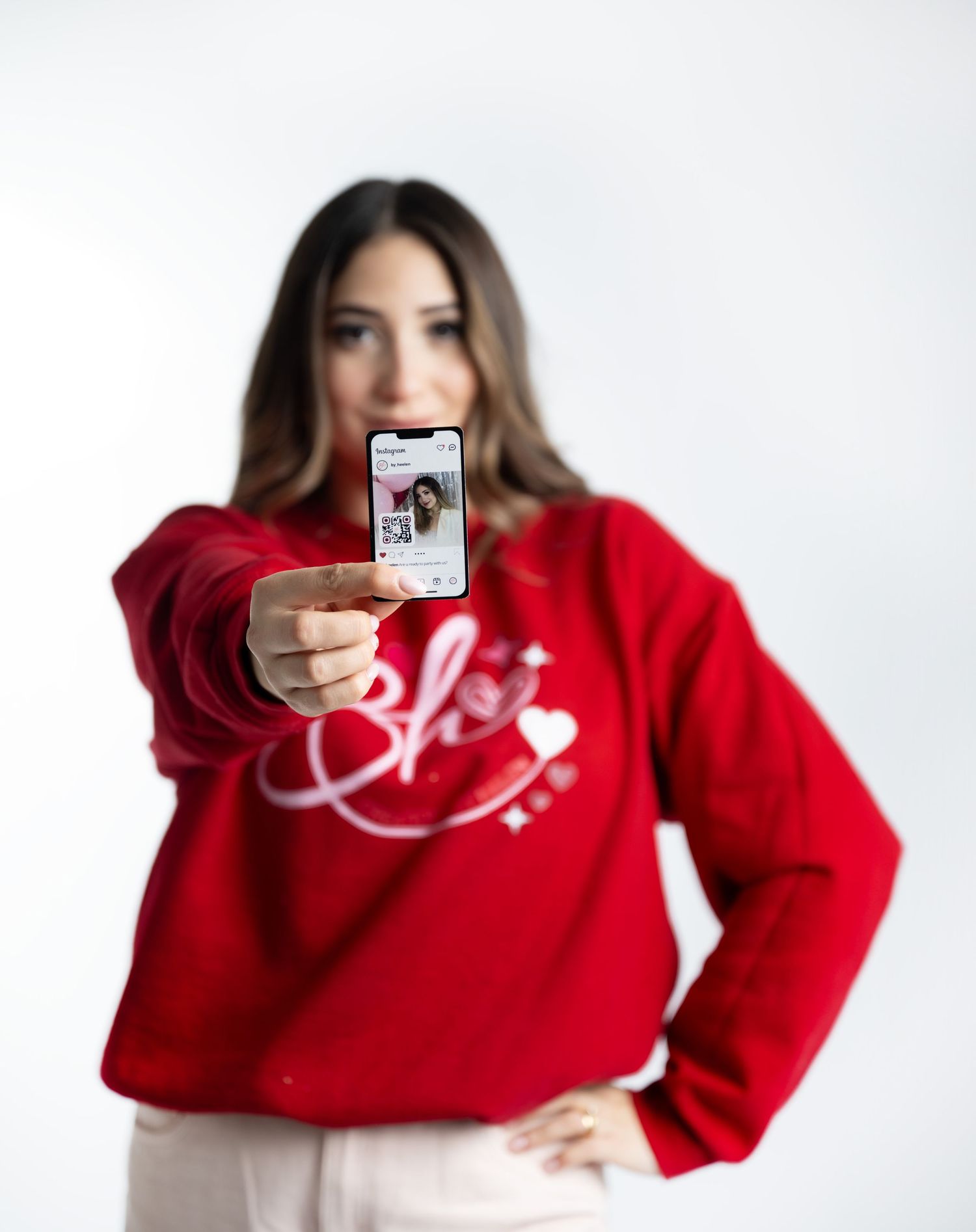 Casual pose in red sweater showing phone screen against white background.