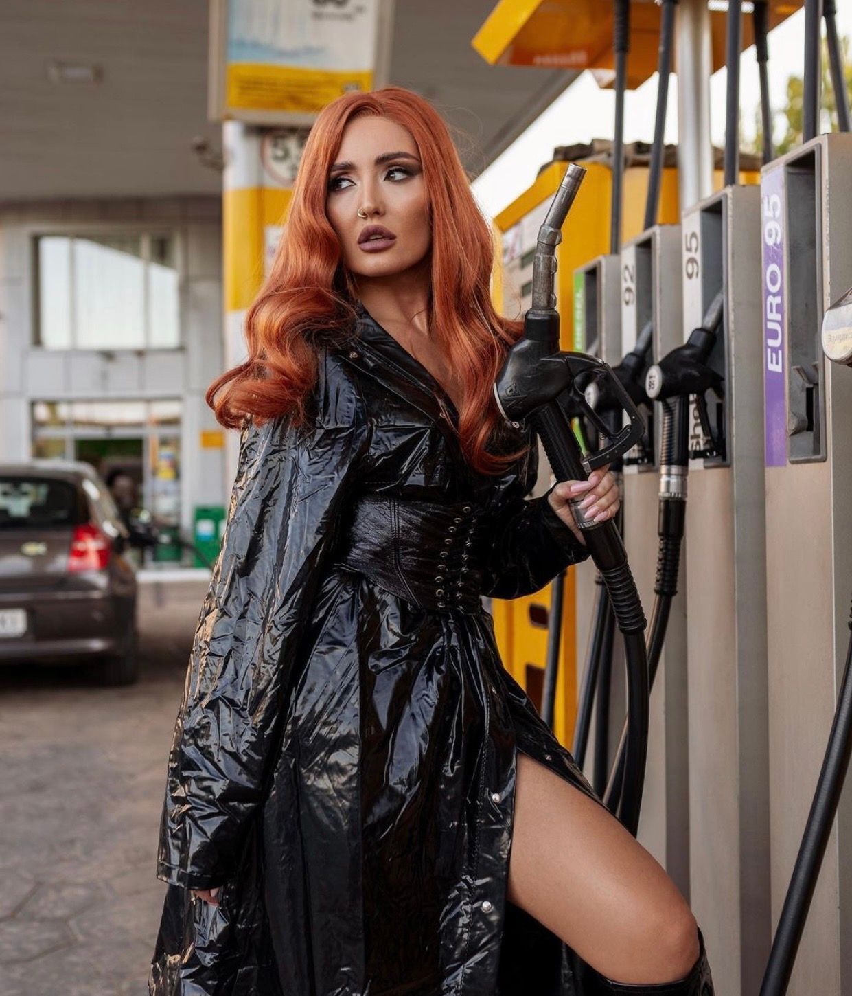 Woman in black vinyl trench coat posing at a gas station with red hair.