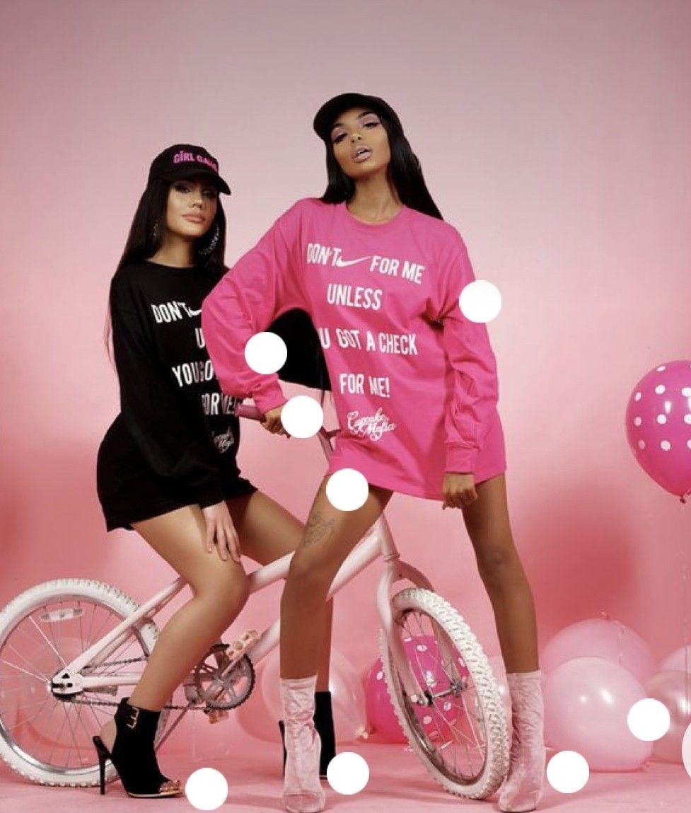 Two women posing in pink and black oversized graphic sweatshirts with a white bicycle and pink balloons in a pink studio.