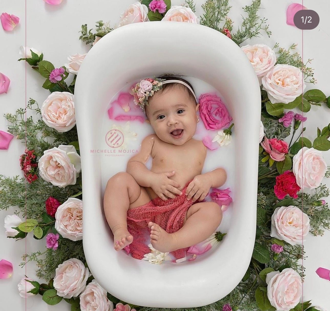 Smiling baby girl in floral headband sitting in white bathtub surrounded by pink roses.