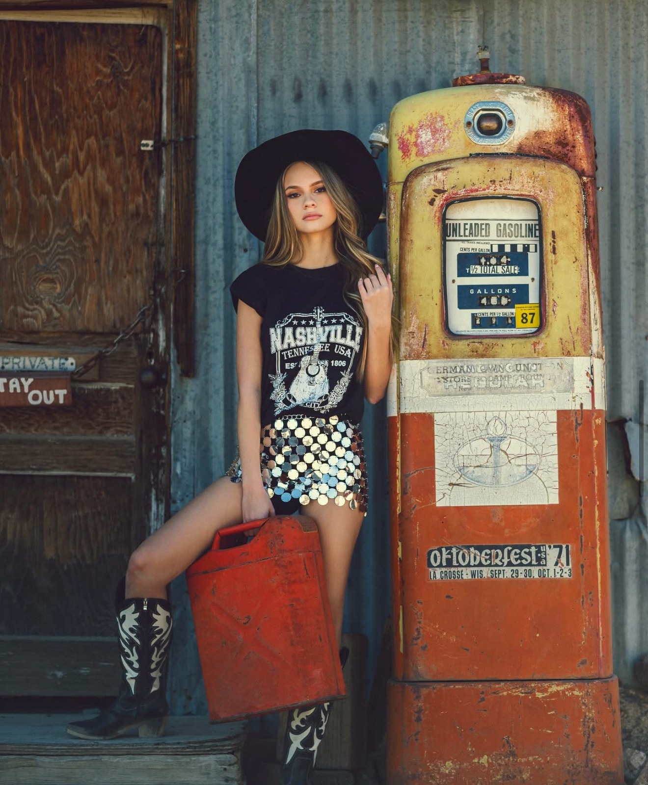 Woman in Nashville tee, sequin shorts, cowboy boots and hat poses by vintage rusty gas pump at weathered building.