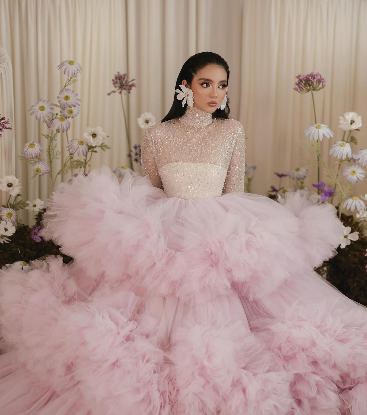 Woman in a voluminous pink tulle ballgown with beaded bodice and floral earrings, surrounded by white daisies.