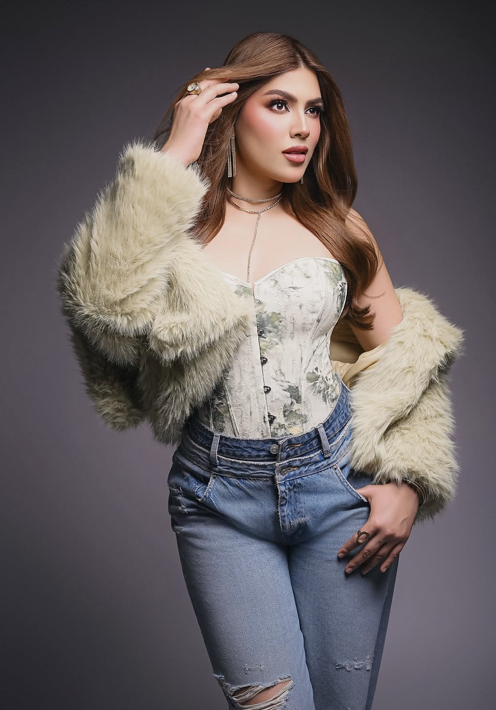 Woman in white floral corset top, cream faux fur jacket, and ripped jeans posing confidently in a studio setting.