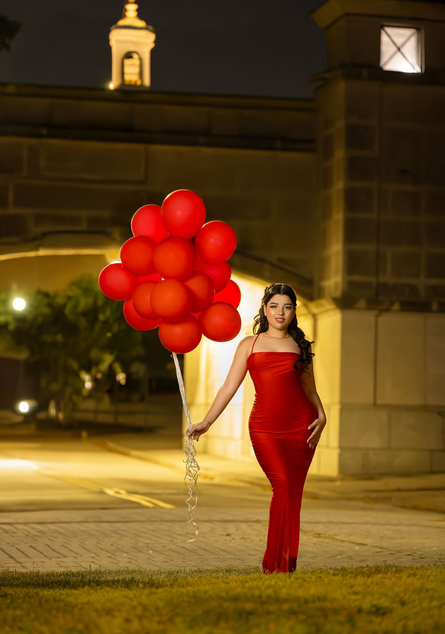A figure in a red dress carries red balloons in a dimly lit urban setting at night.