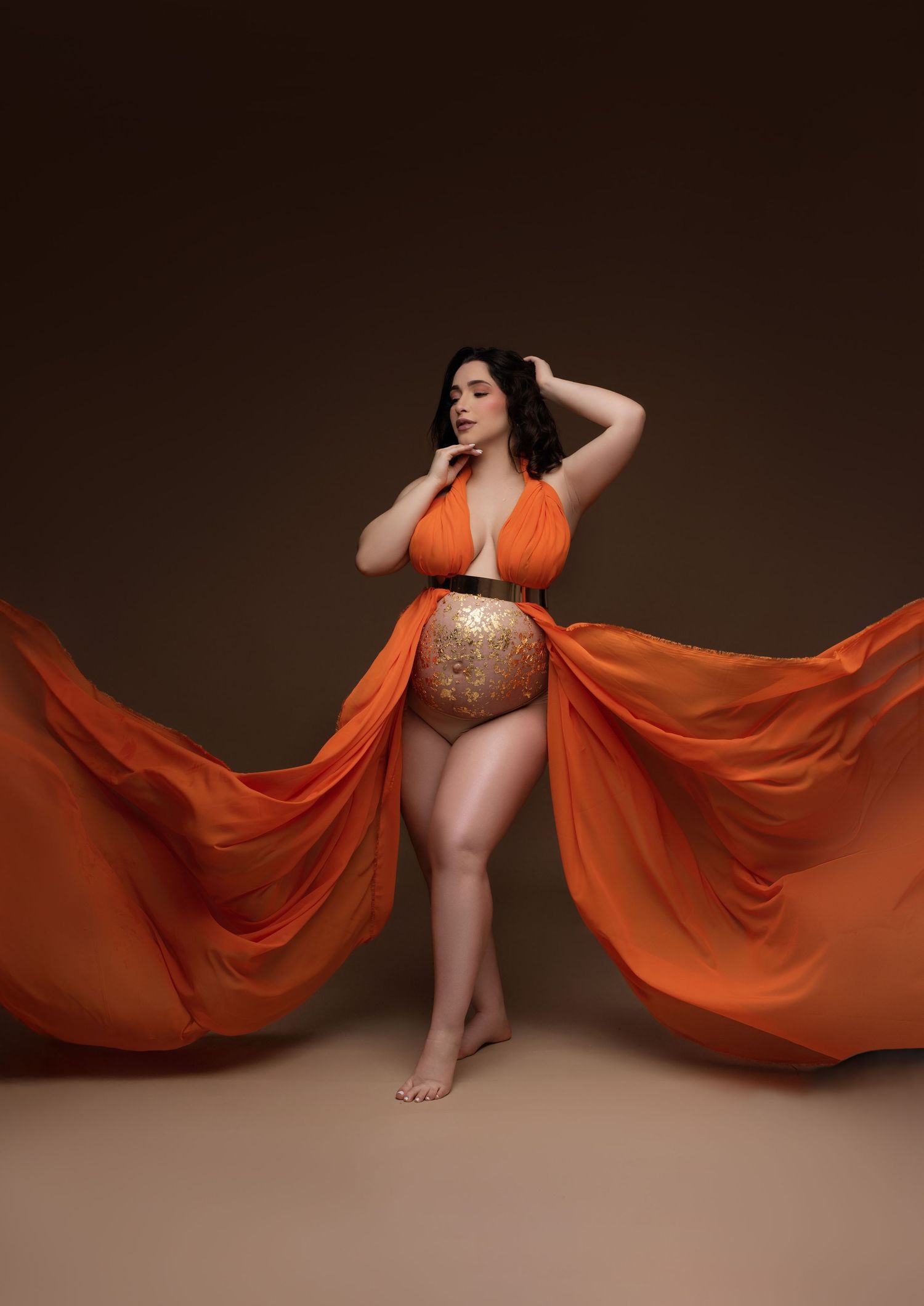 A flowing orange maternity gown with gold belly art creates a dramatic silhouette against a dark brown backdrop.