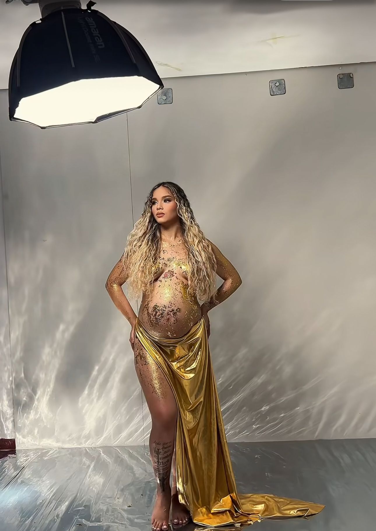 Pregnant woman in gold satin skirt with floral body paint poses under studio lighting for maternity photoshoot.