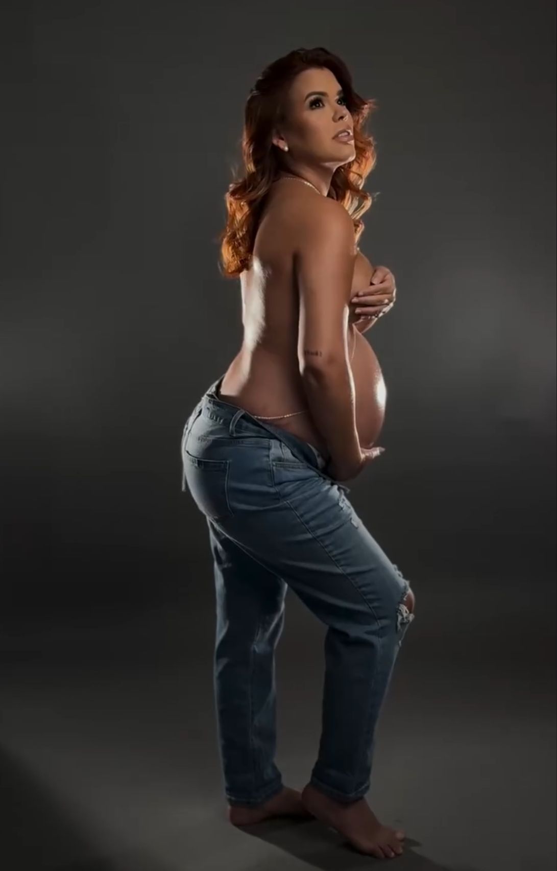 Pregnant woman with red hair posing topless in jeans, cradling baby bump in professional studio photo.