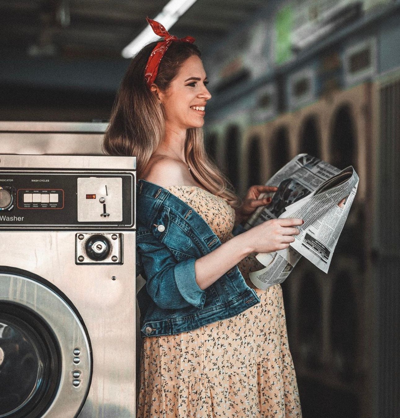 Smiling woman in floral dress and denim jacket reads newspaper at a vintage laundromat with red hair bow.