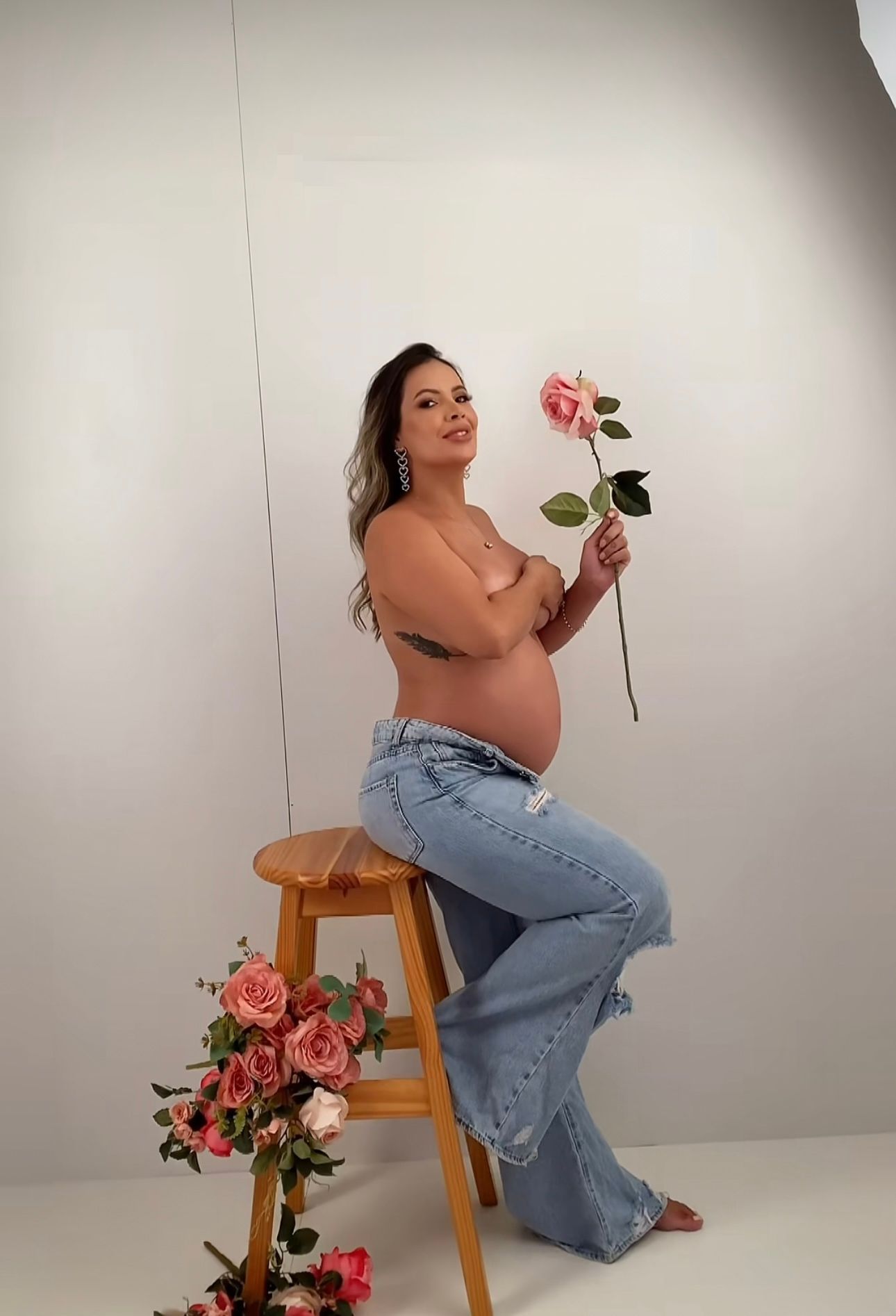 Pregnant woman in jeans holding a pink rose, posing topless for a maternity photo shoot with flowers.