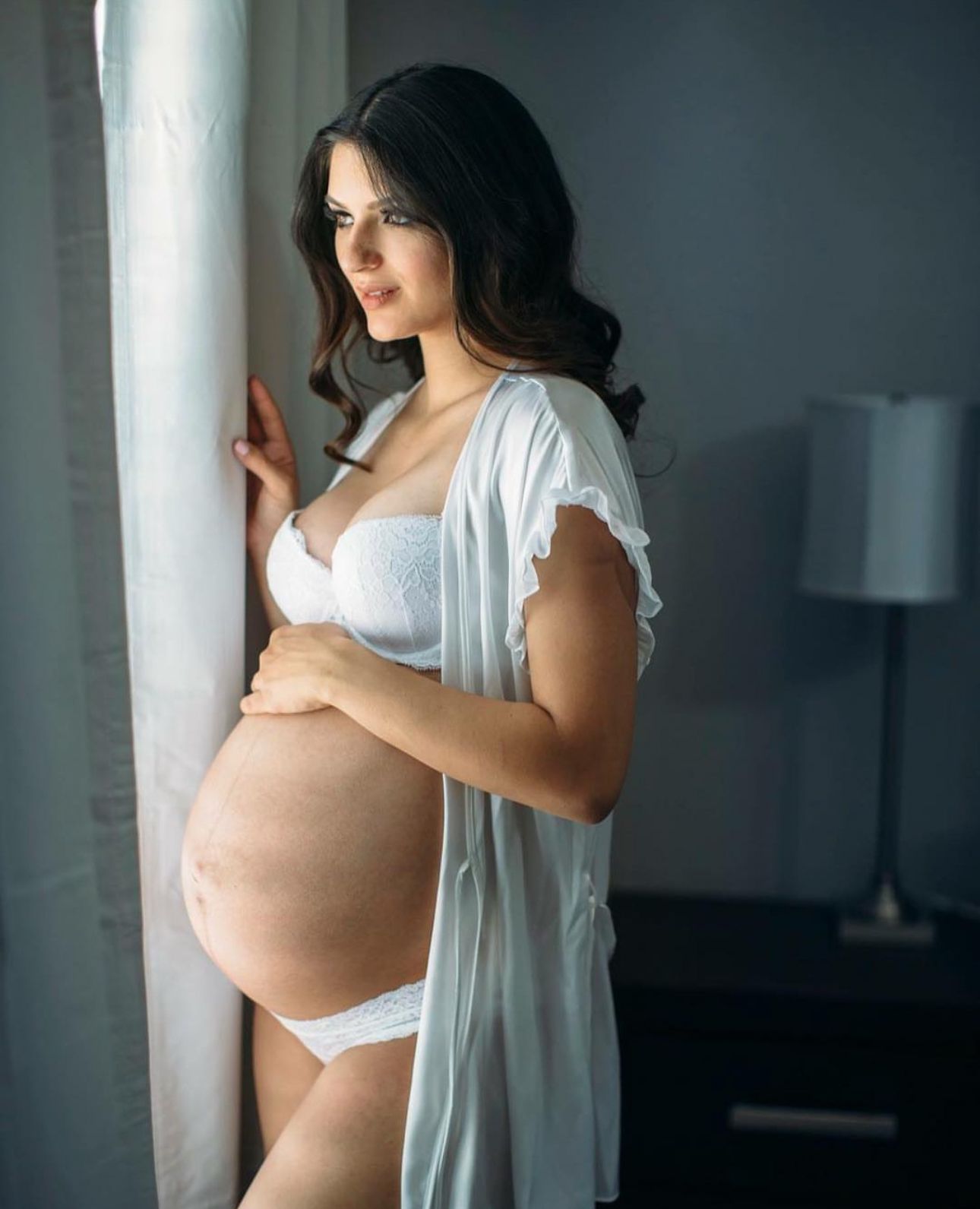 Pregnant woman in white lingerie and sheer robe standing by window cradling baby bump in soft natural light.