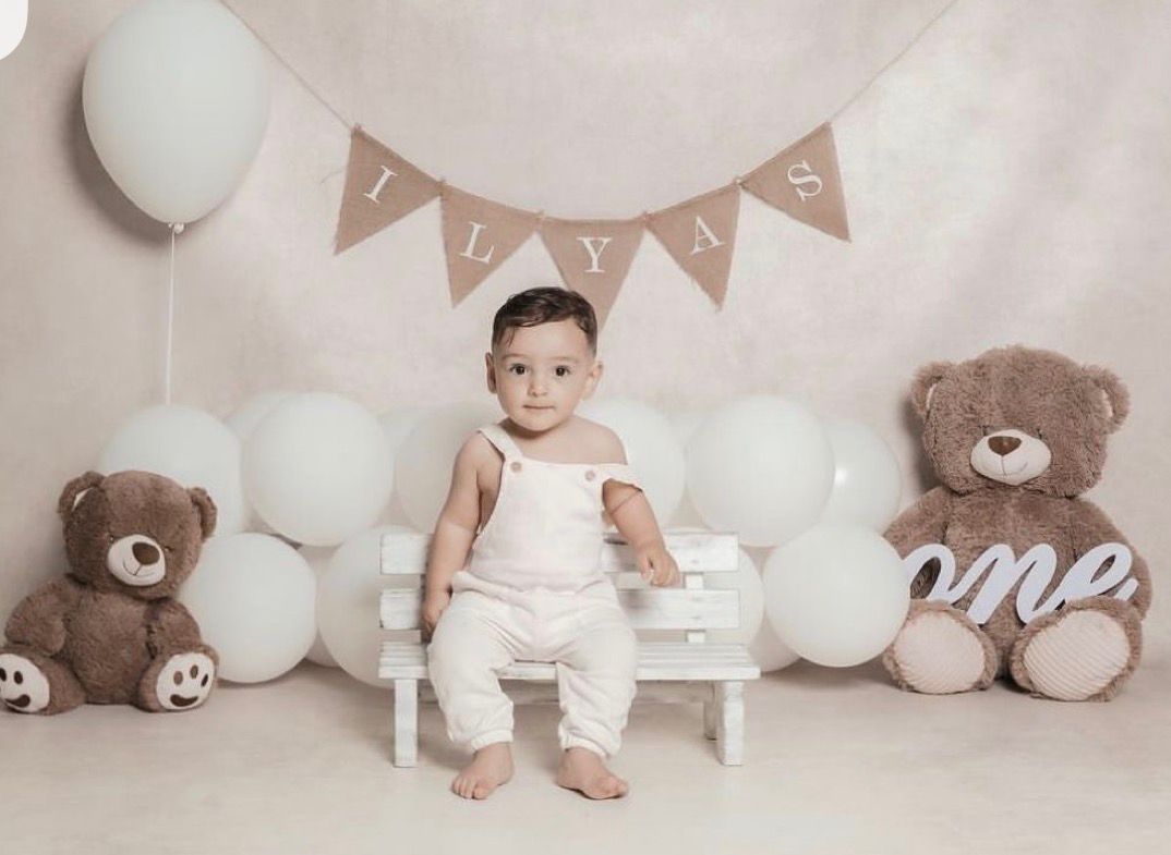 Baby boy Ilyas sits on white bench surrounded by teddy bears and balloons at his first birthday cake smash photo shoot.