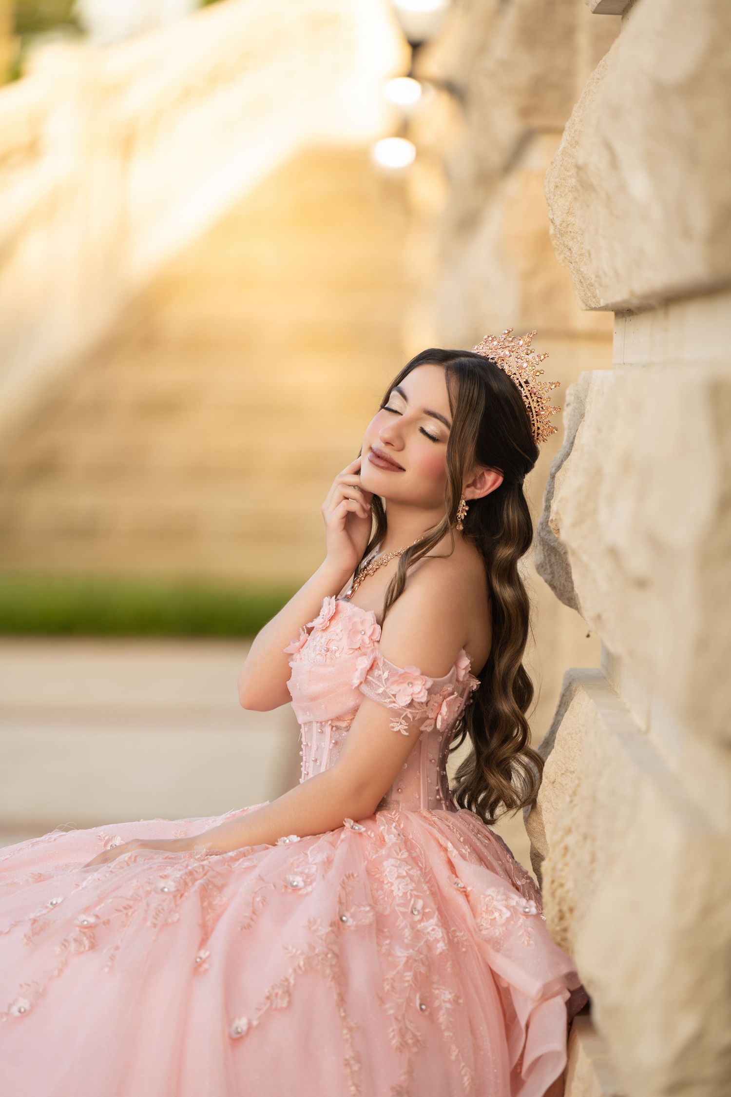 Young woman in pink quinceañera ball gown with floral details and tiara poses gracefully against stone wall.